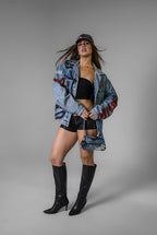 Girl wearing an oversized denim jacket with graffiti and hand painted, black shorts, and black boots on a grey background