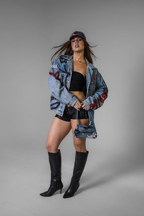 Girl wearing an oversized denim jacket with graffiti and hand painted, black shorts, and black boots on a grey background