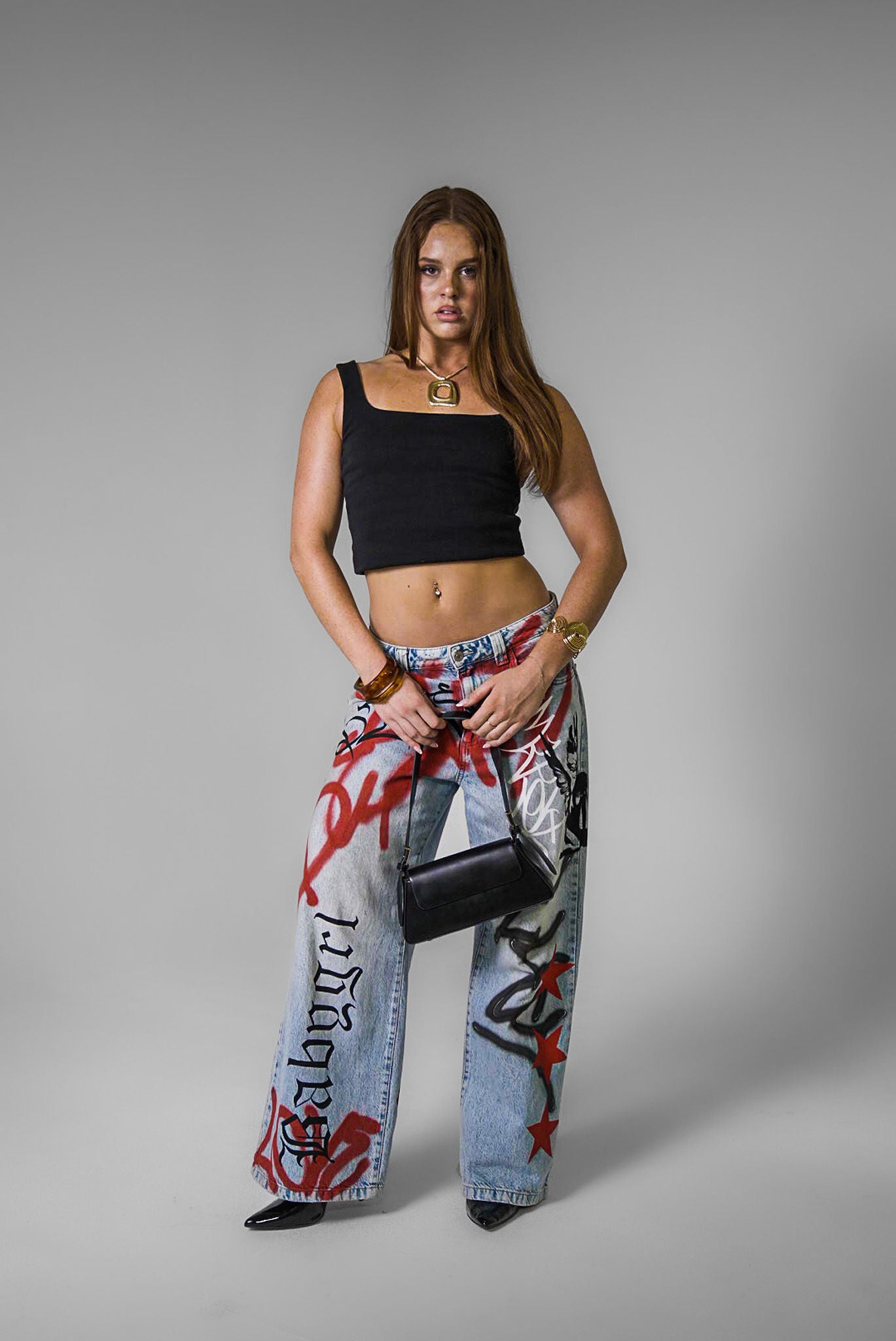 Woman wearing a black crop top and graffiti hand painted denim jeans, holding a black handbag on a grey background.